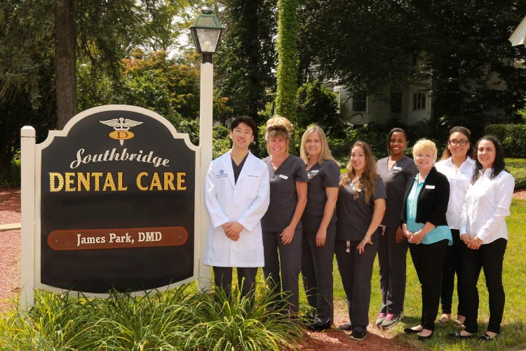 Dr. James Park smiles with his Southbridge Dental Care family by the office sign outside