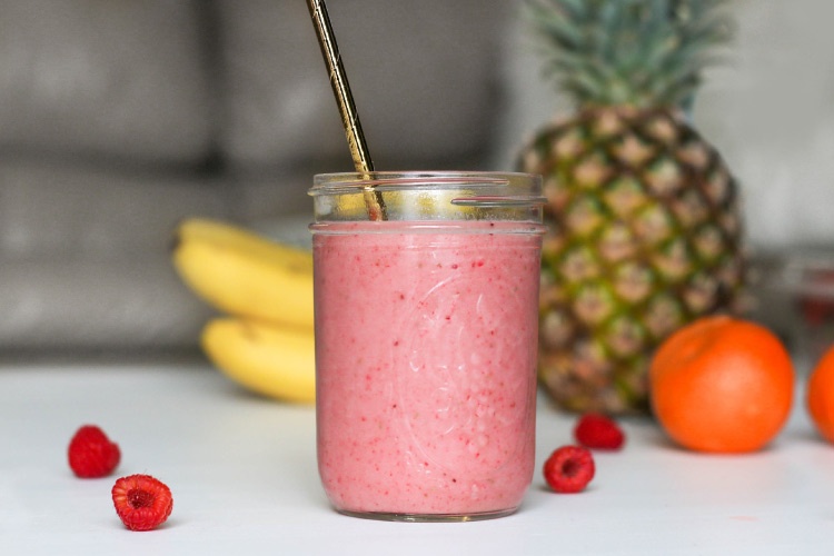 Closeup of a fruit smoothie next to raspberries, an orange, bananas, and a pineapple