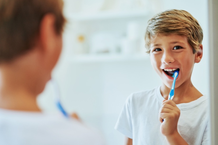 Brunette young boy brushes his teeth with a blue toothbrush while looking in the mirror