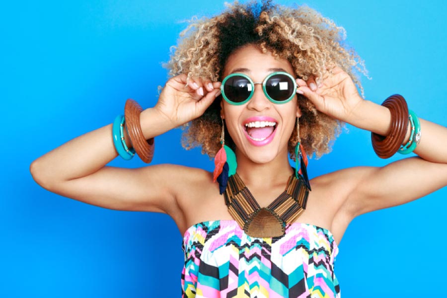 Smiling girl in front of a blue background with big sunglasses, big hair, and funky jewelry