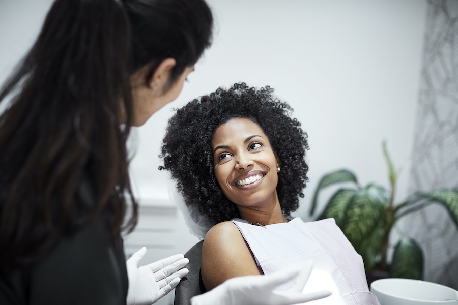 Curly-haired smiling Black woman with a white bib in a dental chair talking to her female dentist